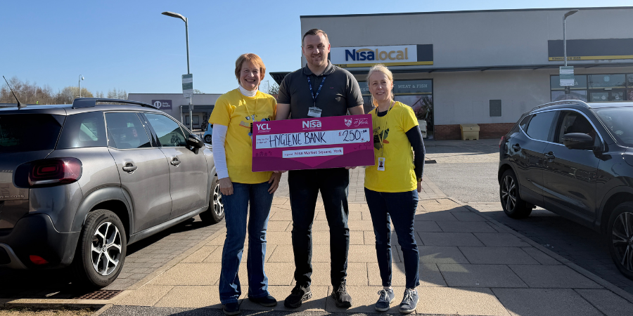 Three people standing outside a Nisa Local store holding a large pink cheque for £250 for The Hygiene Bank. From left to right: Maggie from The Hygiene Bank, Joe from the University of York retail team, and Vicky from The Hygiene Bank.